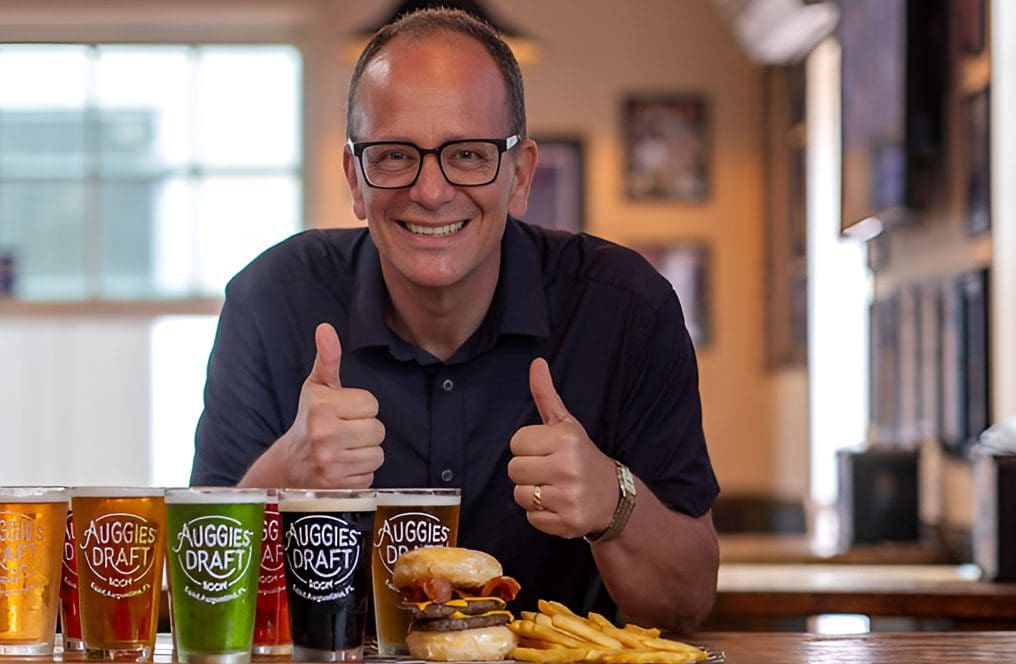 A man sitting at a table with two beers and a burger.